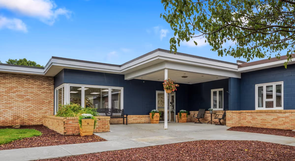 Exterior view of Avantara Clark facility entrance with a covered porch, brick and dark blue walls, large windows, potted plants, and outdoor seating under a partly cloudy sky.