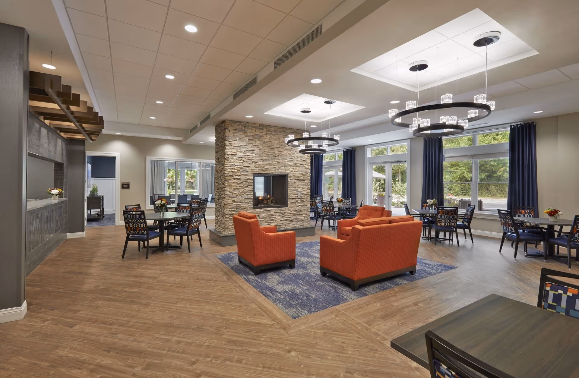 A spacious and modern common area in a senior living facility featuring a central stone fireplace with two circular chandeliers above. The room has several tables with chairs arranged around the perimeter and a seating area with orange armchairs on a blue rug. Large windows with dark curtains allow natural light to fill the space, and the flooring is a combination of wood and carpet.