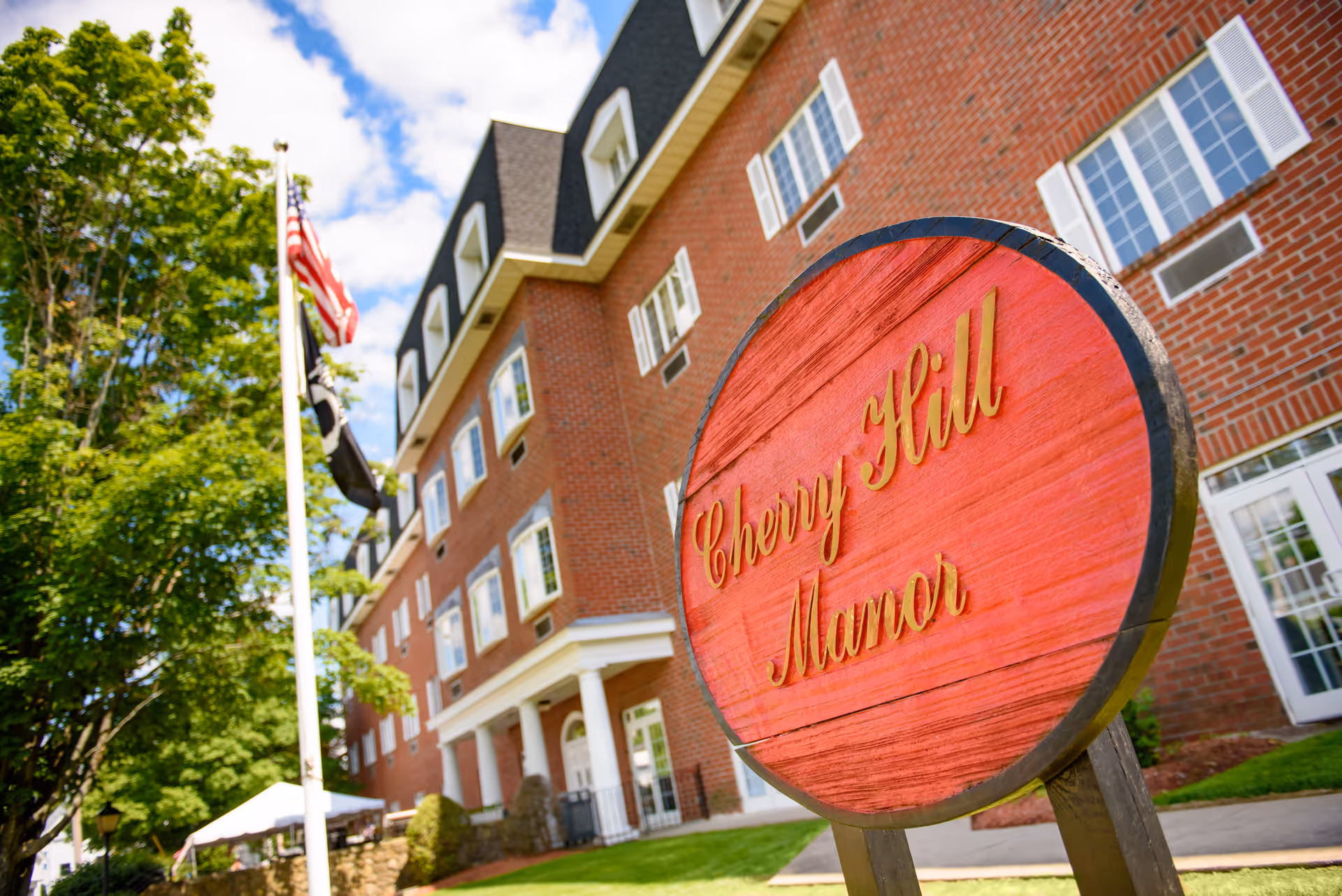 Exterior view of a multi-story brick building with white-trimmed windows and a steep roof. In the foreground, there is a round red wooden sign with gold lettering that reads 'Cherry Hill Manor'. An American flag and another flag are flying on a flagpole near the building, with green trees and a partly cloudy sky in the background.