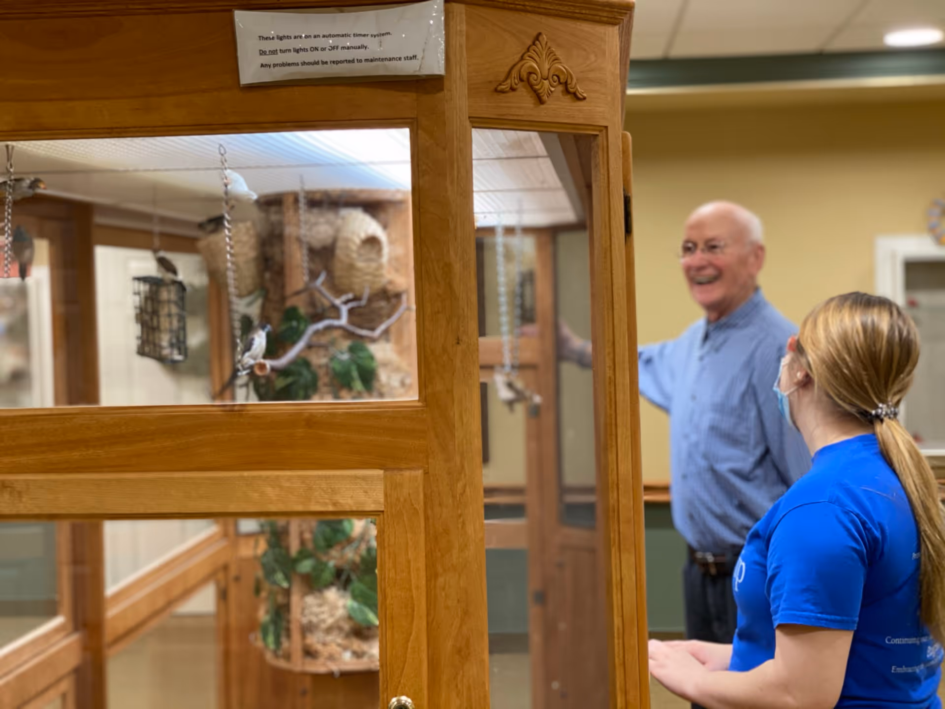 An elderly man and a woman wearing a blue shirt and a face mask are standing near a wooden enclosure with glass panels that contains bird feeders and decorative birdhouses inside a senior living facility.