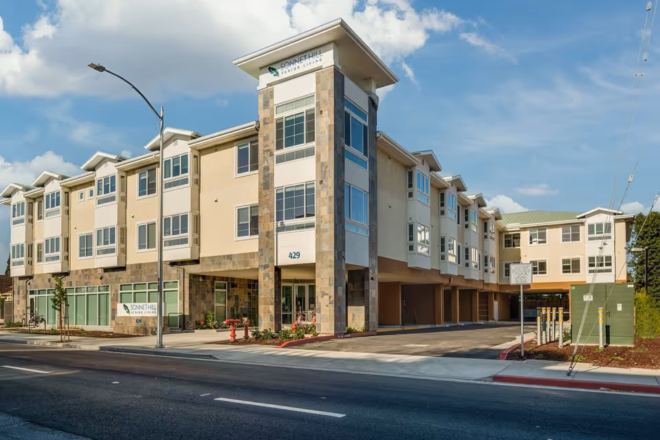 Exterior view of Sonnet Hill Senior Living building, a modern three-story structure with large windows, stone accents, and a covered parking area. The building is situated along a street with a sidewalk, streetlamp, and some landscaping. The sky is partly cloudy.