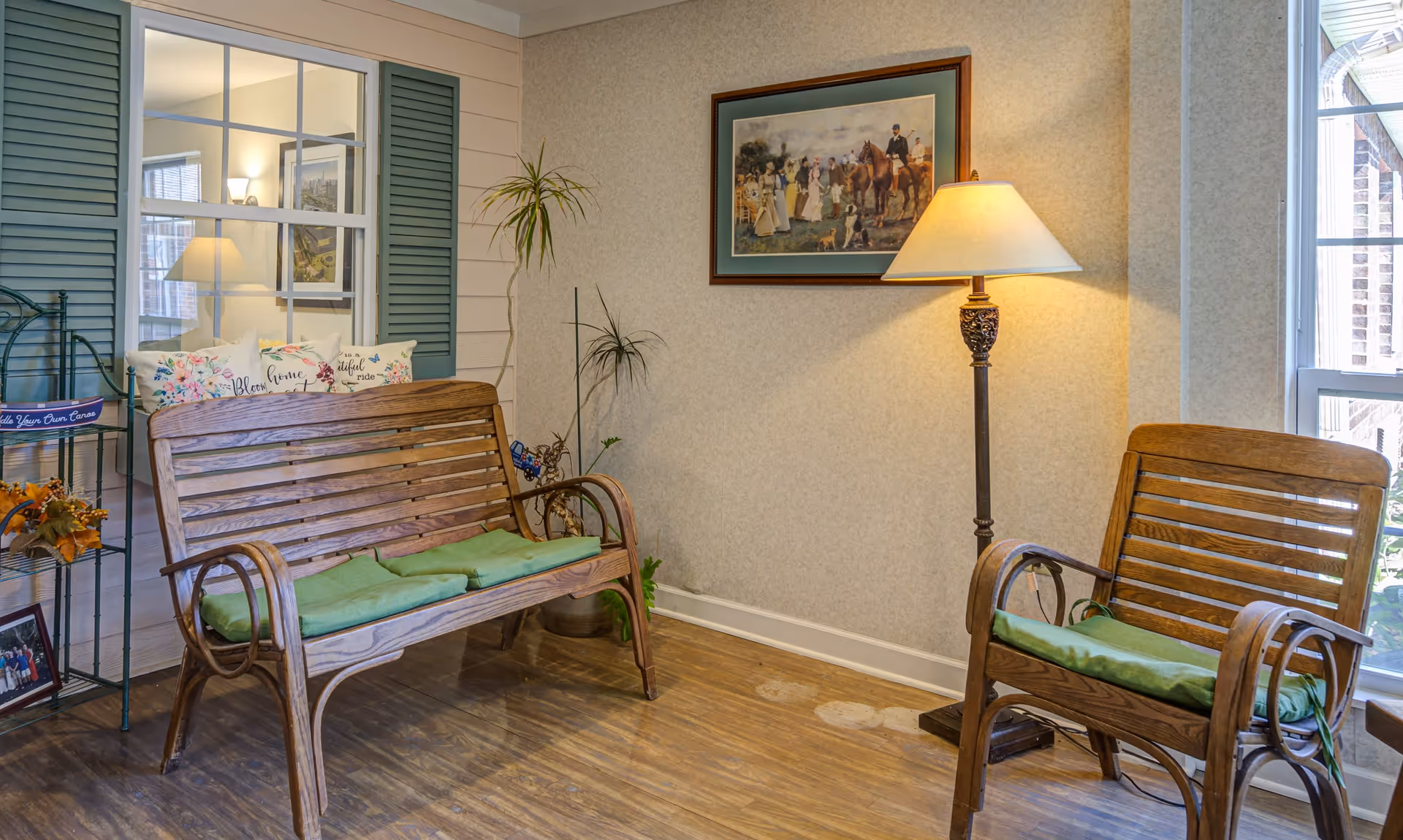 A cozy seating area with wooden bench and chair, both with green cushions. A tall floor lamp with a beige shade is lit beside the chair. There is a framed painting of people and horses on the wall above the lamp. A metal plant stand with decorative items and pillows is placed near the bench. A window with green shutters and a large window letting in natural light are visible.