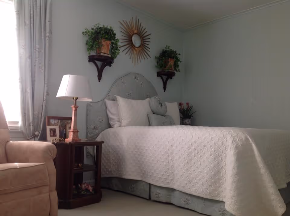 A softly lit bedroom with a neatly made bed covered by a white quilt, an upholstered headboard, bedside table with a lamp, and an armchair.
