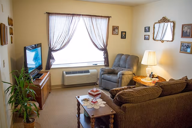 Cozy living room with a sofa, recliner, TV, coffee table and lamp by a curtained window.