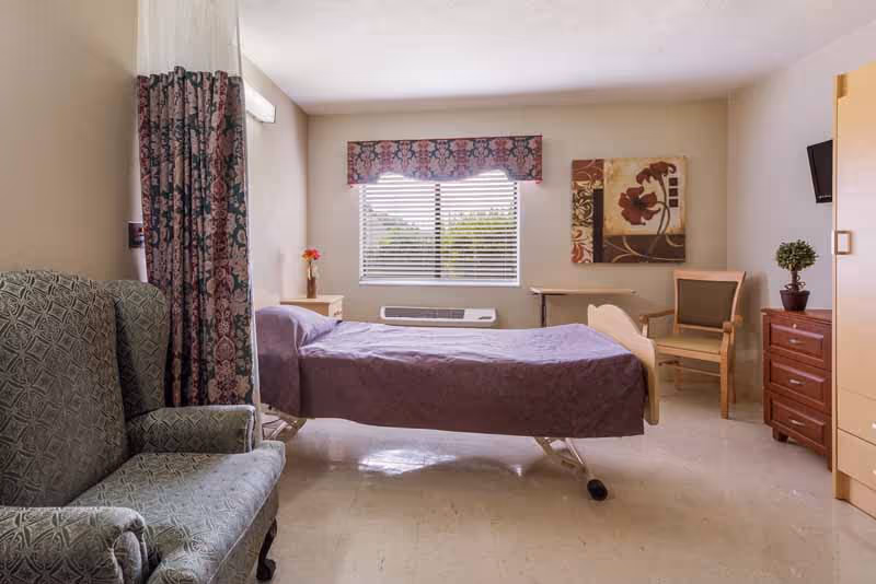 A simple, clean bedroom in a healthcare facility featuring a single hospital bed with a purple bedspread, a green upholstered armchair, a wooden dresser with a small potted plant, a wooden chair, a small table, a window with blinds and a floral valance, and a floral wall painting.