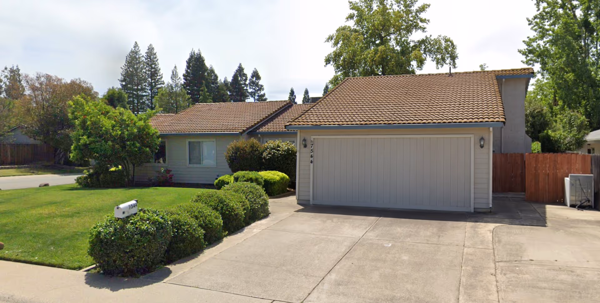 Front view of a single-story suburban house with a tiled roof, two-car garage, driveway, and well-kept lawn and shrubs.