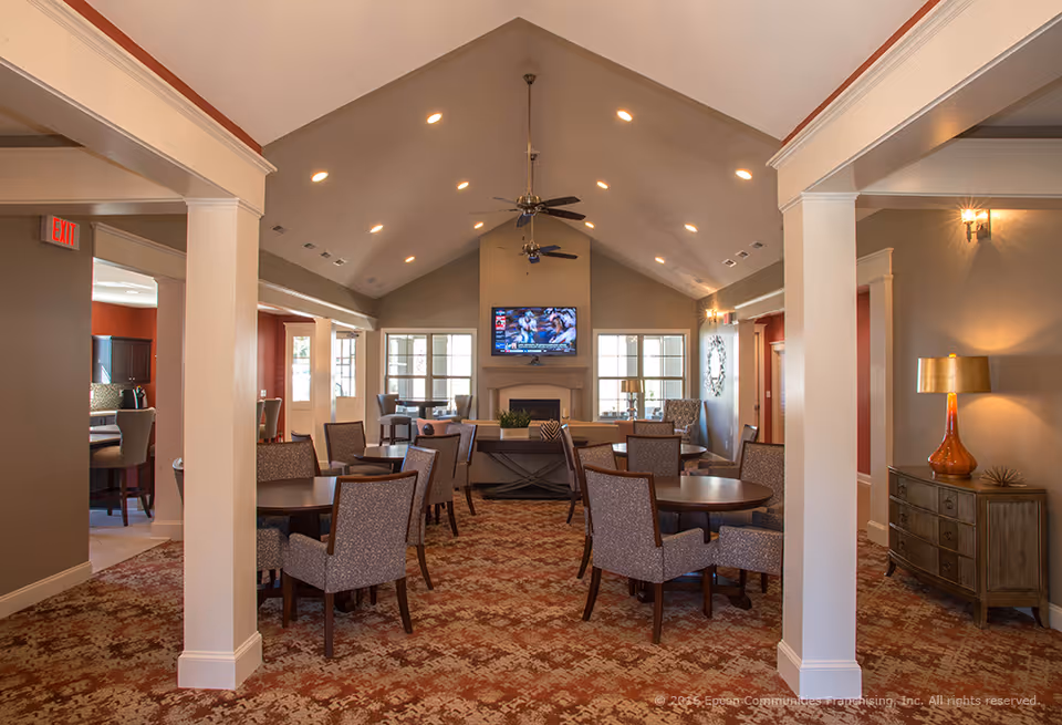 Community lounge with round dining tables and upholstered chairs facing a wall-mounted TV and fireplace under a vaulted ceiling.