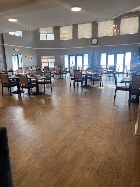 Spacious dining room with multiple tables and chairs arranged neatly on a wooden floor. Large windows and glass doors allow natural light to fill the room. A decorated Christmas tree is visible near the windows, and chandeliers hang from the ceiling. A clock is mounted on the wall above the tree.