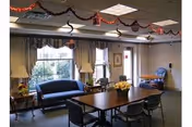 A senior living common room with a central rectangular table and chairs, a sofa by windows, lamps, and decorative garlands hanging from the ceiling.
