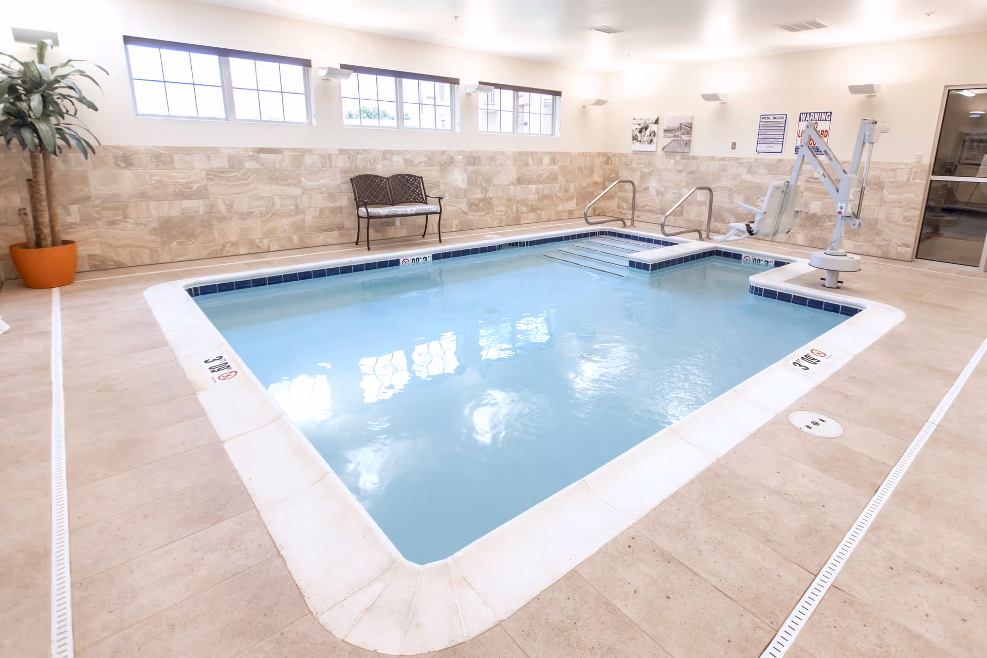 Indoor swimming pool with clear blue water, surrounded by beige tiled flooring and walls with stone accents. There is a metal bench with cushions against the wall, a potted plant in the corner, pool safety signs, and a pool lift for accessibility.
