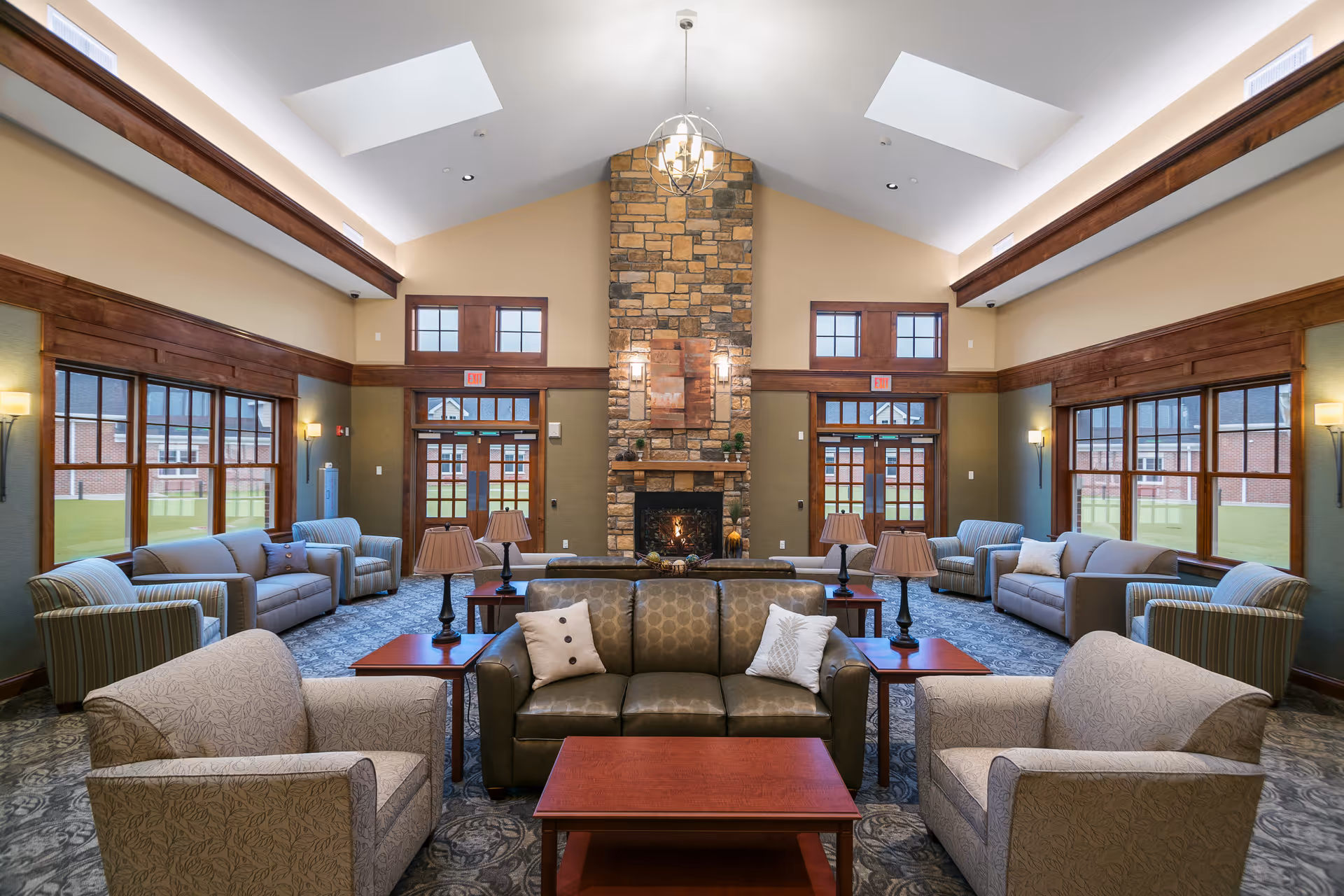 Spacious and well-lit living room with high vaulted ceilings and skylights. The room features a large stone fireplace centered on the far wall, surrounded by multiple seating areas including sofas and armchairs arranged around wooden coffee tables. Large windows line the walls, allowing natural light to fill the space. The room has a warm and inviting atmosphere with neutral tones and wooden trim accents.