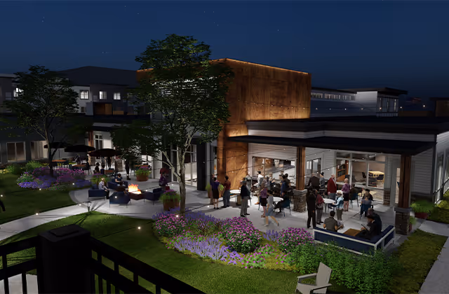 Nighttime view of an outdoor patio area at a senior living facility with people gathered around a fire pit and seated on outdoor furniture. The building is modern with large windows and warm lighting. There are landscaped flower beds and trees surrounding the patio.