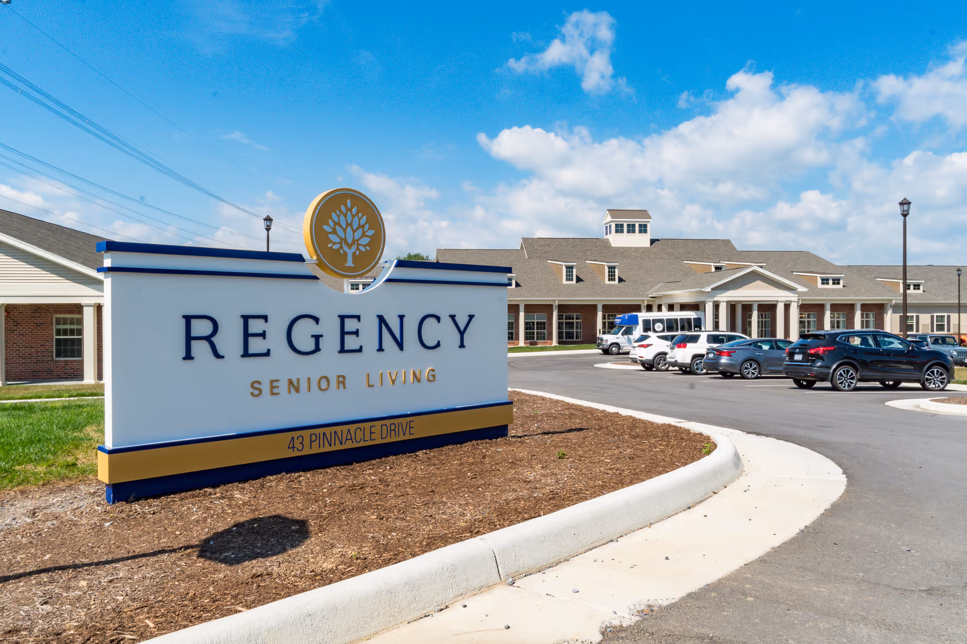 Front exterior of the Regency Senior Living building with a large property sign, parking lot and cars under a blue sky.