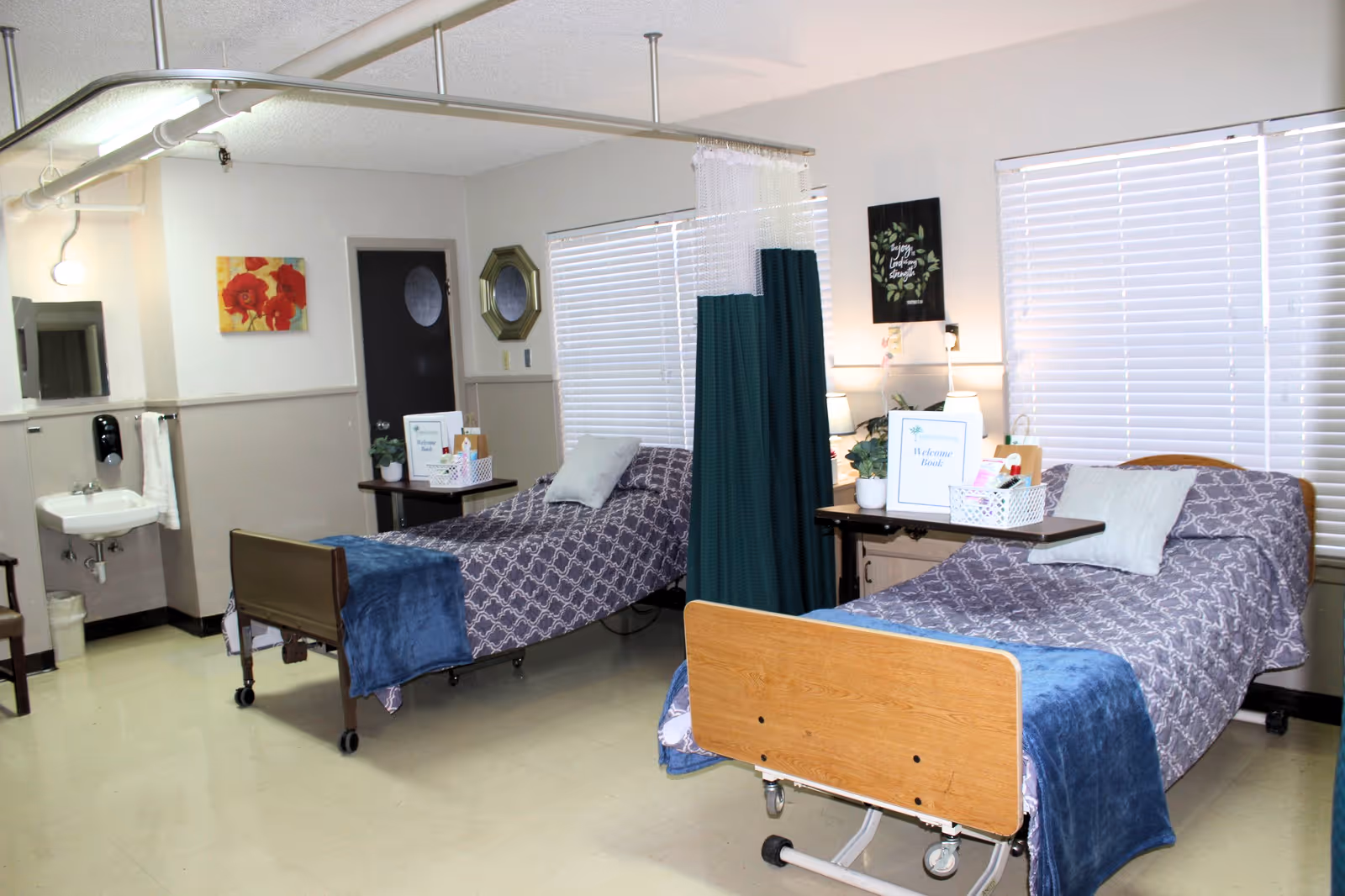 A nursing facility room with two hospital beds separated by a green privacy curtain. Each bed is made with patterned bedding and a blue blanket at the foot. There are bedside tables with lamps, plants, and welcome signs. The room has large windows with blinds, a wall-mounted sink with a towel, and simple wall decor including a painting and a mirror.