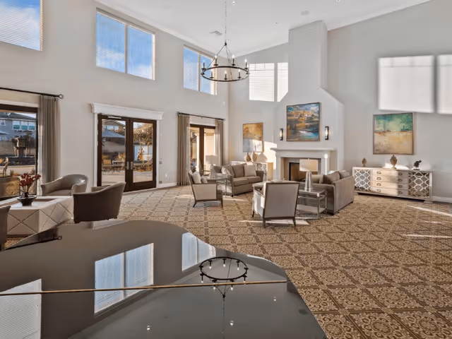 Spacious and well-lit living room area with high ceilings, large windows, and glass doors leading outside. The room features a patterned carpet, a chandelier, a fireplace with artwork above it, multiple seating arrangements including sofas and armchairs, a sideboard with decorative items, and a grand piano in the foreground.