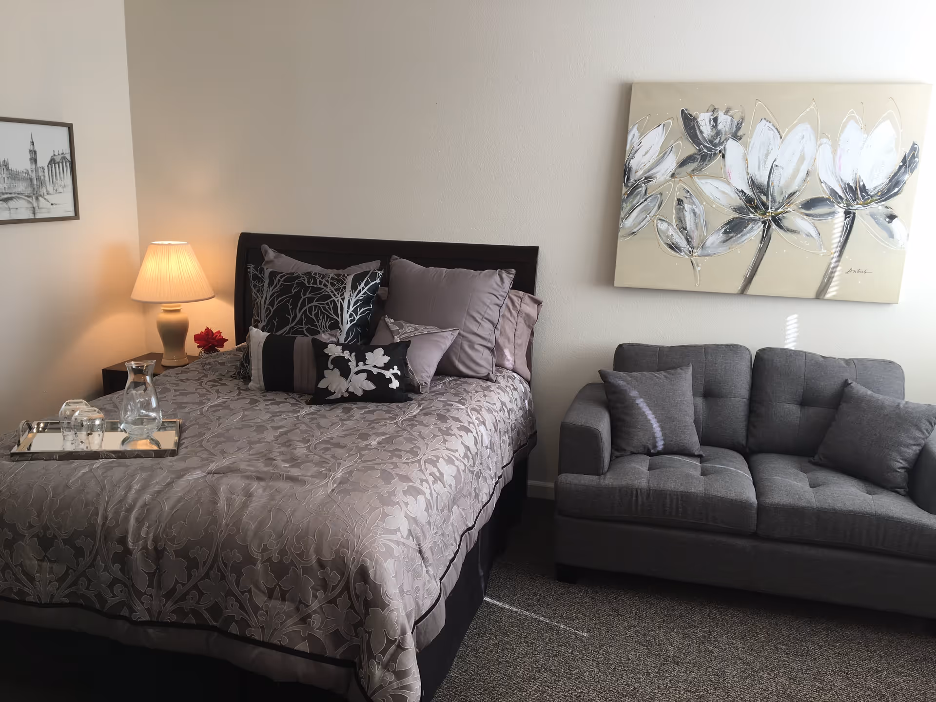 A cozy bedroom featuring a neatly made bed with multiple decorative pillows and a patterned bedspread. Next to the bed is a nightstand with a lamp and a small red flower. A gray cushioned loveseat with two pillows is positioned against the wall, beneath a large floral painting. The room has neutral-colored walls and carpeted flooring.