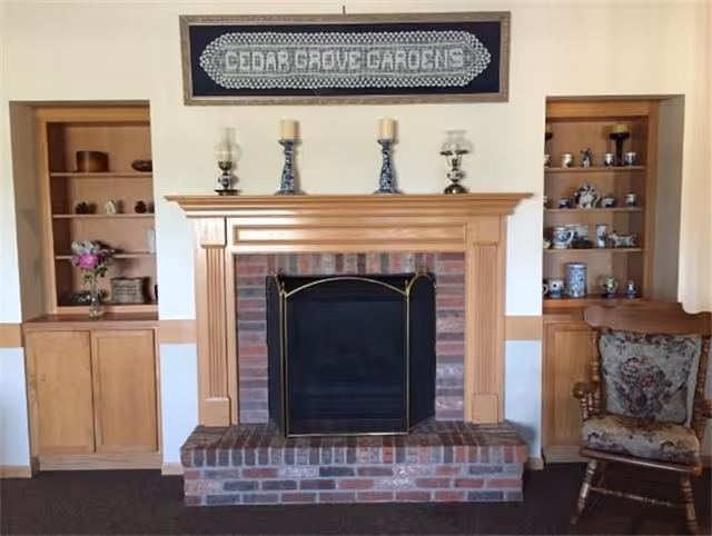 Brick fireplace with a wooden mantel flanked by built-in shelves, decorative items, a rocking chair, and a 'Cedar Grove Gardens' sign above.