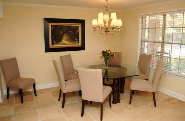 A dining room with a glass-top table surrounded by six beige upholstered chairs. A chandelier with multiple lights hangs above the table. A vase with flowers is placed at the center of the table. There is a large window with blinds on the right side and a framed picture of a tree-lined path on the wall.