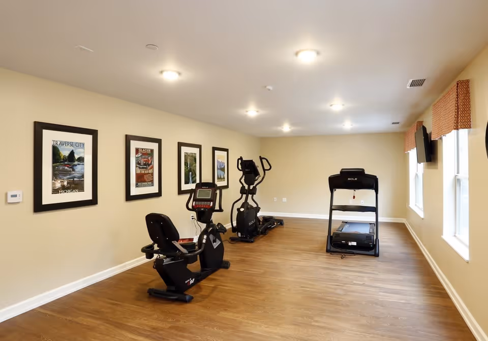 A small fitness room with wooden flooring and beige walls. The room contains three pieces of exercise equipment: a recumbent bike, an elliptical machine, and a treadmill. Four framed pictures hang on the left wall, and two windows with patterned valances are on the right wall. A flat-screen TV is mounted on the right wall near the windows.