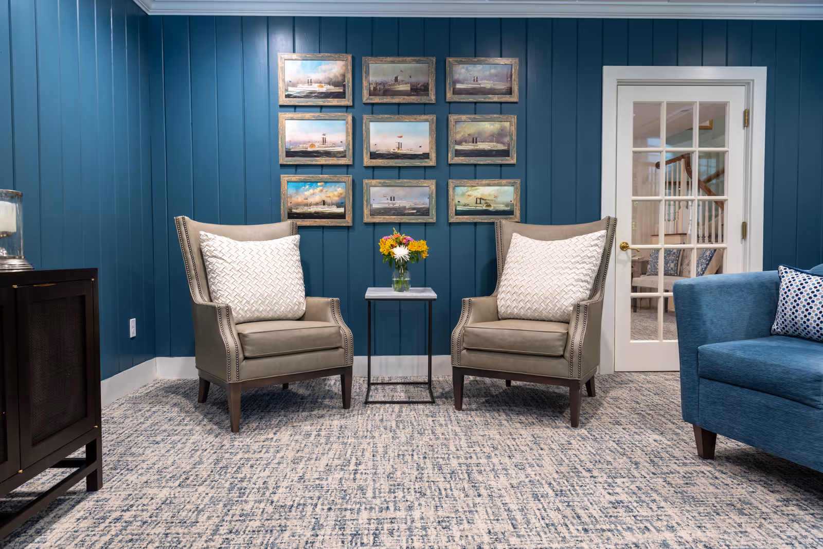 A cozy sitting area with two beige armchairs featuring white patterned cushions, separated by a small table holding a vase of flowers. The background wall is painted teal and decorated with nine framed pictures arranged in a grid. To the right, there is a blue sofa with a patterned pillow and a white door with glass panes leading to another room.