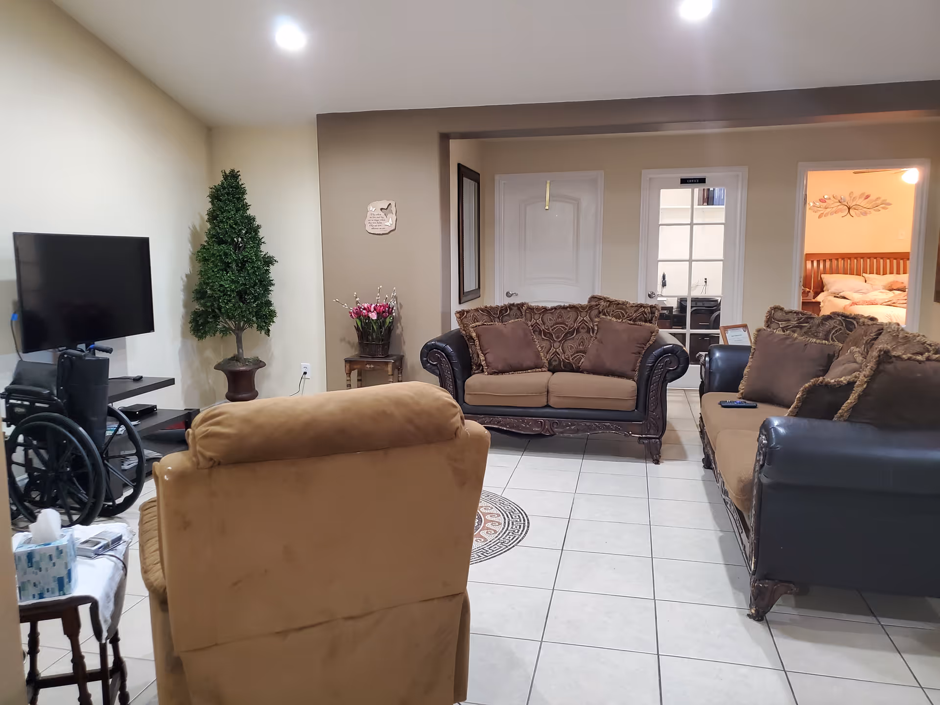 A cozy living room with beige tiled floor, featuring a brown armchair in the foreground, two brown and black sofas with patterned cushions, a flat-screen TV on a stand, a wheelchair, a small table with a tissue box, a potted artificial tree, and a small table with a flower arrangement. There are two doors in the background, one leading to a bedroom with a wooden bed and wall decoration.