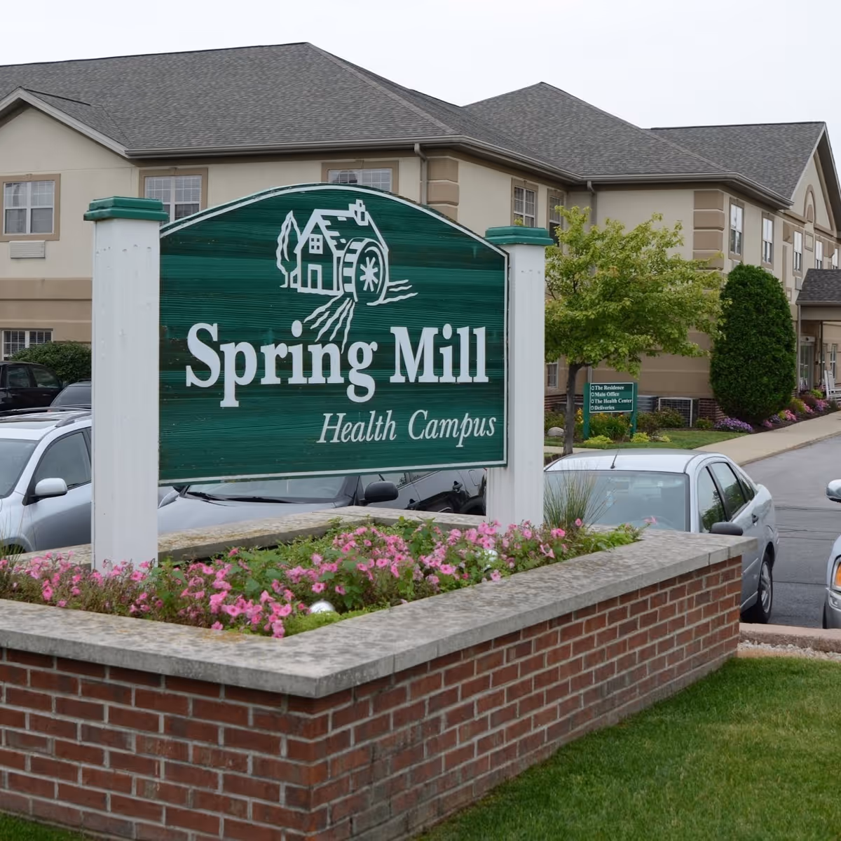 A green wooden sign with white text and a graphic of a watermill, reading 'Spring Mill Health Campus,' is mounted on white posts above a brick planter filled with pink flowers. Behind the sign is a beige two-story building with multiple windows, a tree, and parked cars along a driveway.