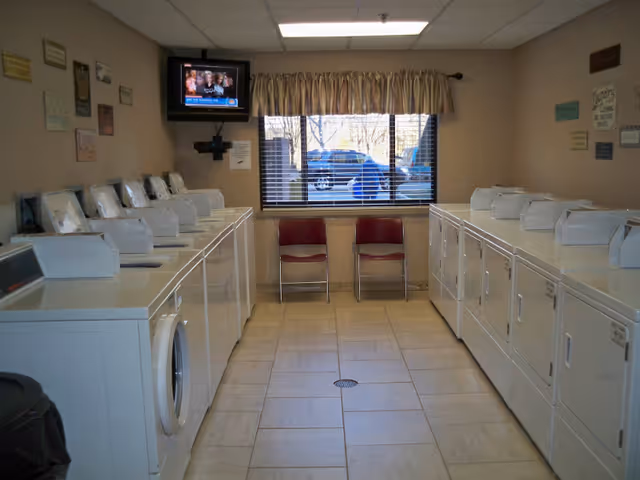 Laundry room with two rows of white washing machines and dryers facing each other. There are two red chairs against the wall under a window with blinds and a beige curtain. A small television is mounted on the wall in the corner above the machines. Various signs and plaques are displayed on the beige walls.