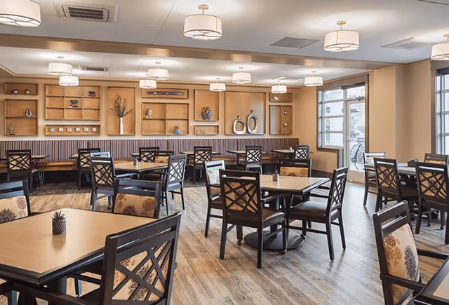A bright and spacious dining room with multiple wooden tables and chairs arranged neatly. The room features light wood flooring, beige walls, built-in wooden shelves with decorative items, and large windows allowing natural light to enter. Ceiling lights provide additional illumination.