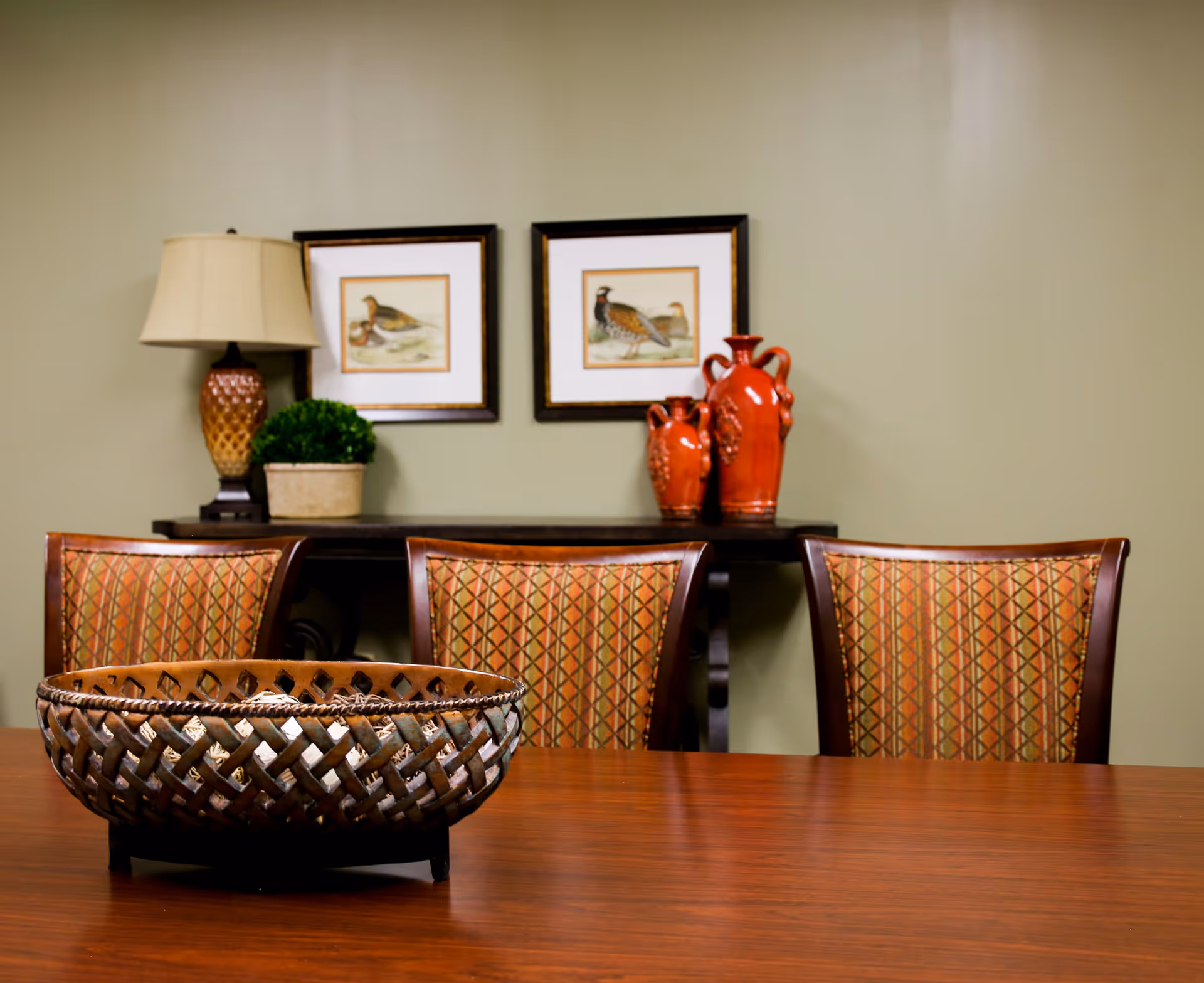 Wood dining table with a woven decorative bowl, three upholstered chairs, and a side table holding a lamp, framed bird prints, and red vases against a green wall.