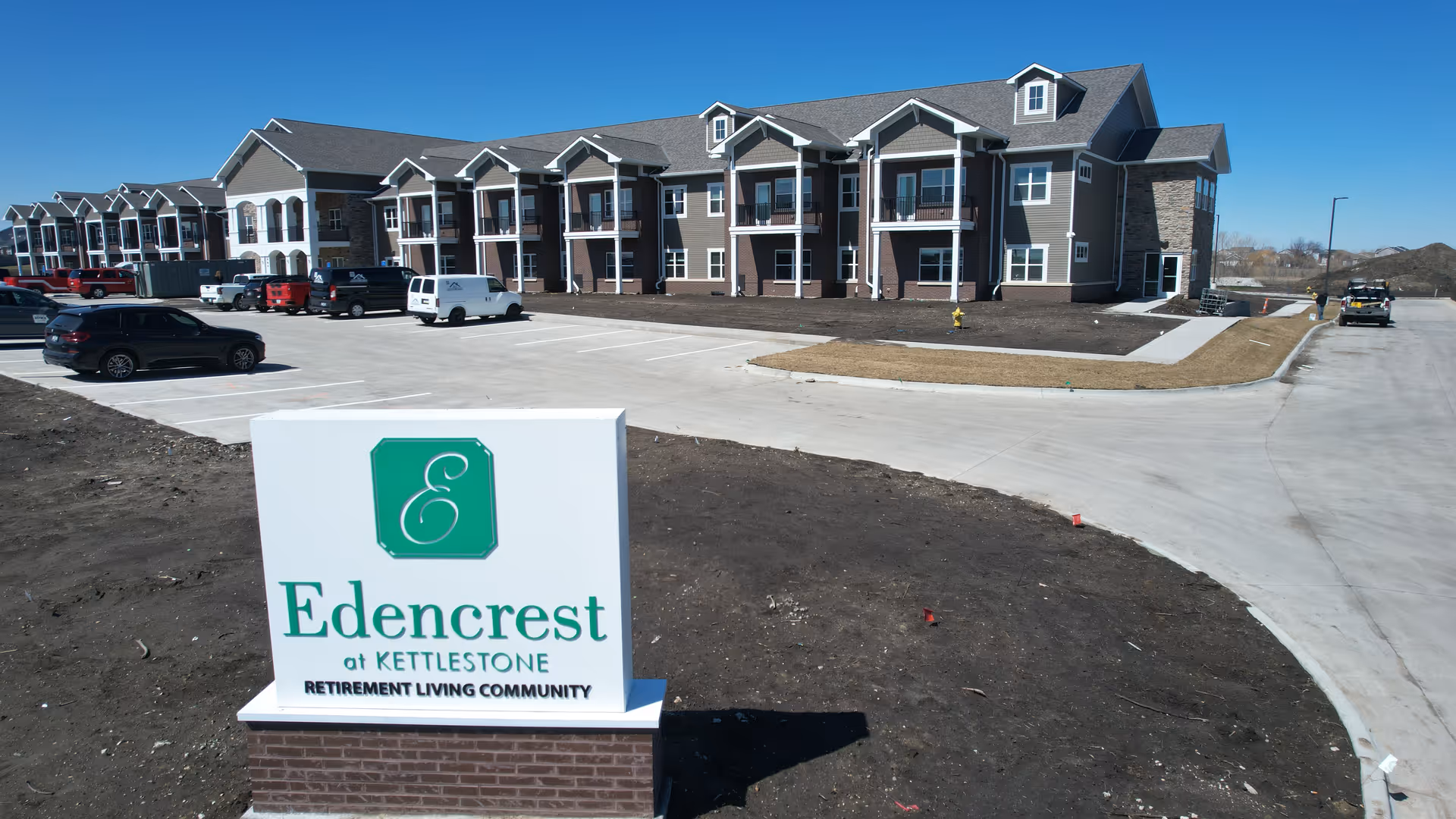 Exterior view of Edencrest at Kettlestone retirement living community showing a large multi-story building with balconies and a parking lot with several vehicles under a clear blue sky.