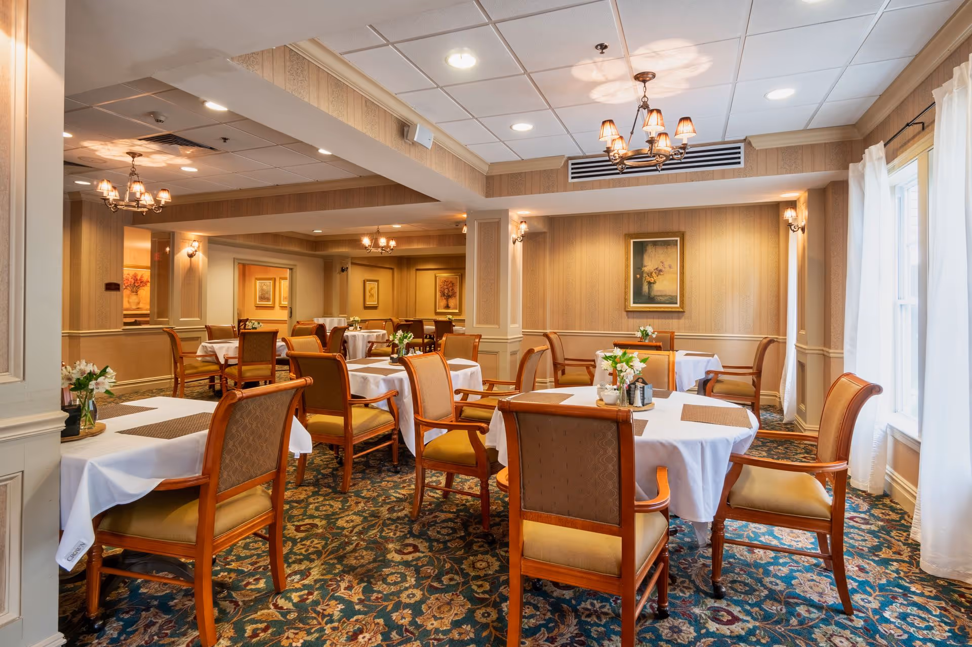 A warmly lit dining room with multiple tables covered in white tablecloths and brown placemats. Each table is surrounded by wooden chairs with cushioned seats and backs. The room has patterned carpet flooring, beige wallpaper, framed artwork on the walls, and chandeliers hanging from the ceiling. Natural light enters through large windows with sheer white curtains on the right side.