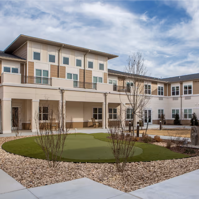 Courtyard of a two-story cream-colored senior living building featuring a small artificial putting green, bare landscaping, and covered walkways with seating under a cloudy sky.