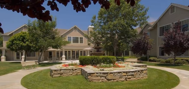Sunlit courtyard with a raised stone flower bed, benches, trees and a two-story senior living building under a clear blue sky.
