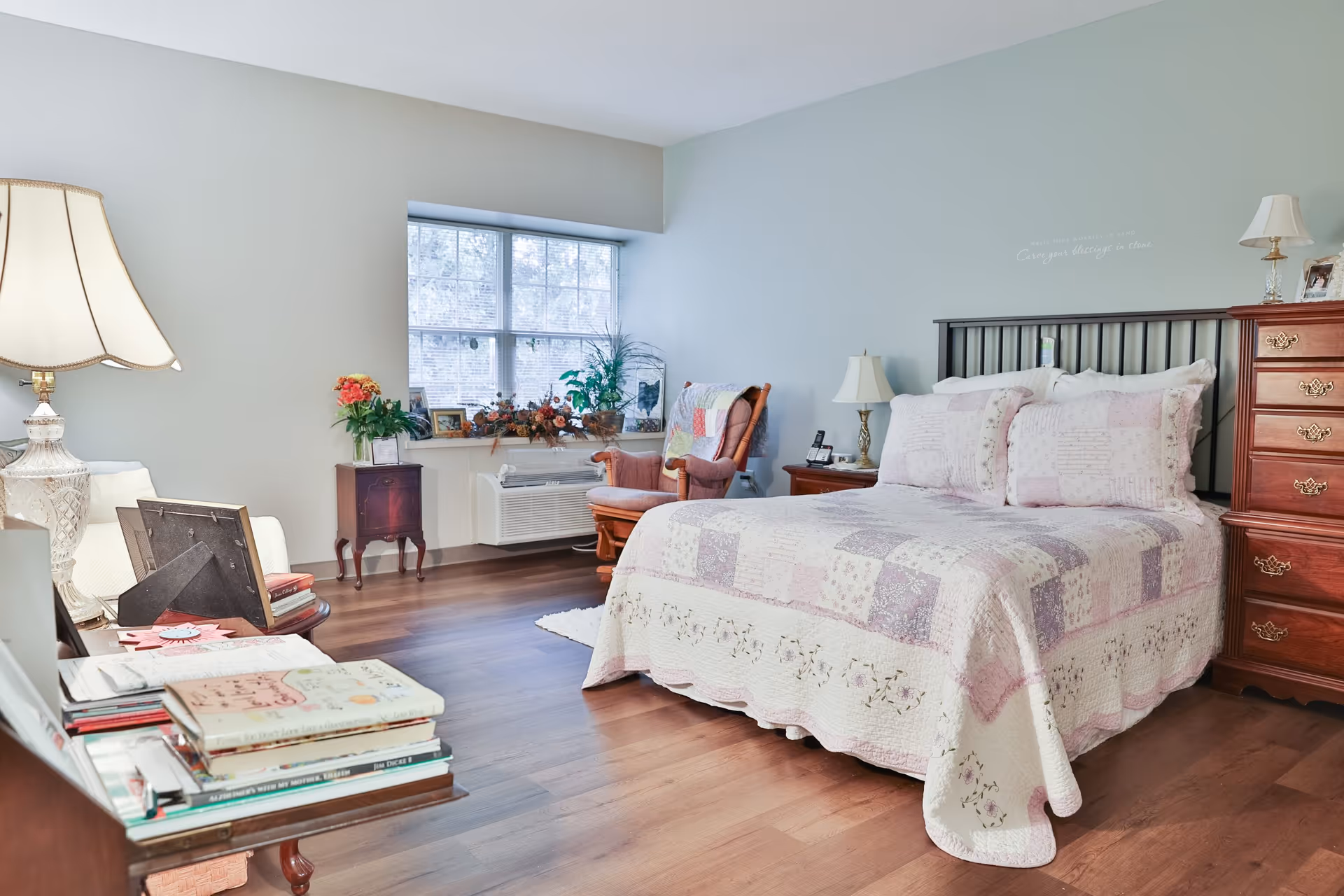 A cozy bedroom in a senior living facility featuring a bed with a patchwork quilt and matching pillows, a wooden dresser with a lamp and framed photos, a comfortable armchair with a quilt draped over it, a window with plants on the sill, and a side table with a lamp and books.