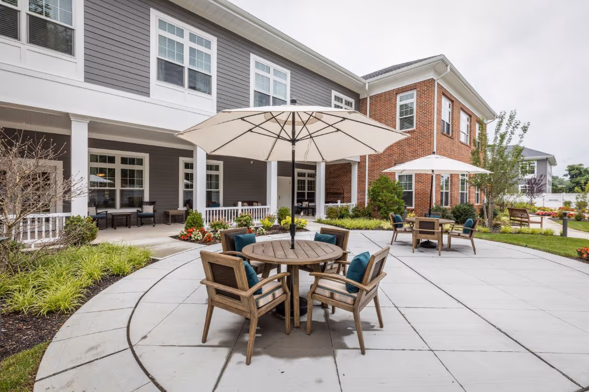 Outdoor patio area at Sunrise of Huntington featuring round wooden tables with umbrellas and cushioned chairs, surrounded by landscaped greenery and a two-story building with gray and red brick exterior walls.