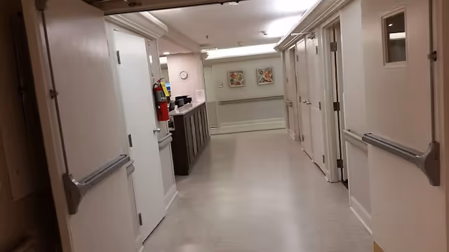 A clean, well-lit hallway inside a facility with multiple closed doors on both sides. There is a fire extinguisher mounted on the left wall near a counter area, and two framed floral pictures are visible on the far wall at the end of the corridor.
