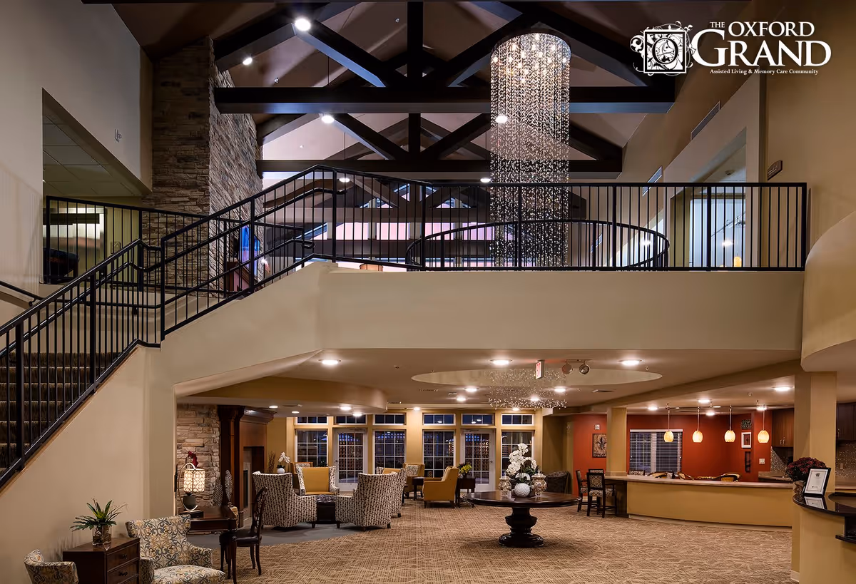 Spacious and elegant interior of The Oxford Grand featuring a high ceiling with exposed dark wooden beams, a large modern chandelier, a staircase with black railings, comfortable seating areas with patterned and yellow chairs, a round table with decorative vases and flowers, and a reception or dining counter with pendant lights.