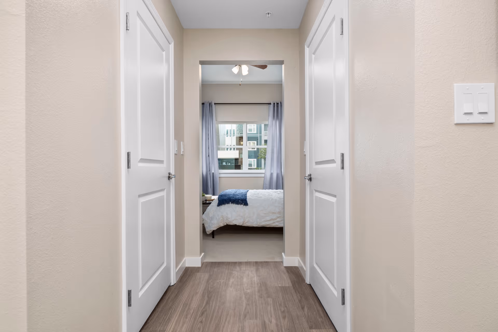 View down a hallway with wood flooring and beige walls, featuring two closed white doors on either side. At the end of the hallway is a bedroom with a bed covered in a white comforter and a blue throw blanket, a window with light blue curtains, and a ceiling fan above.