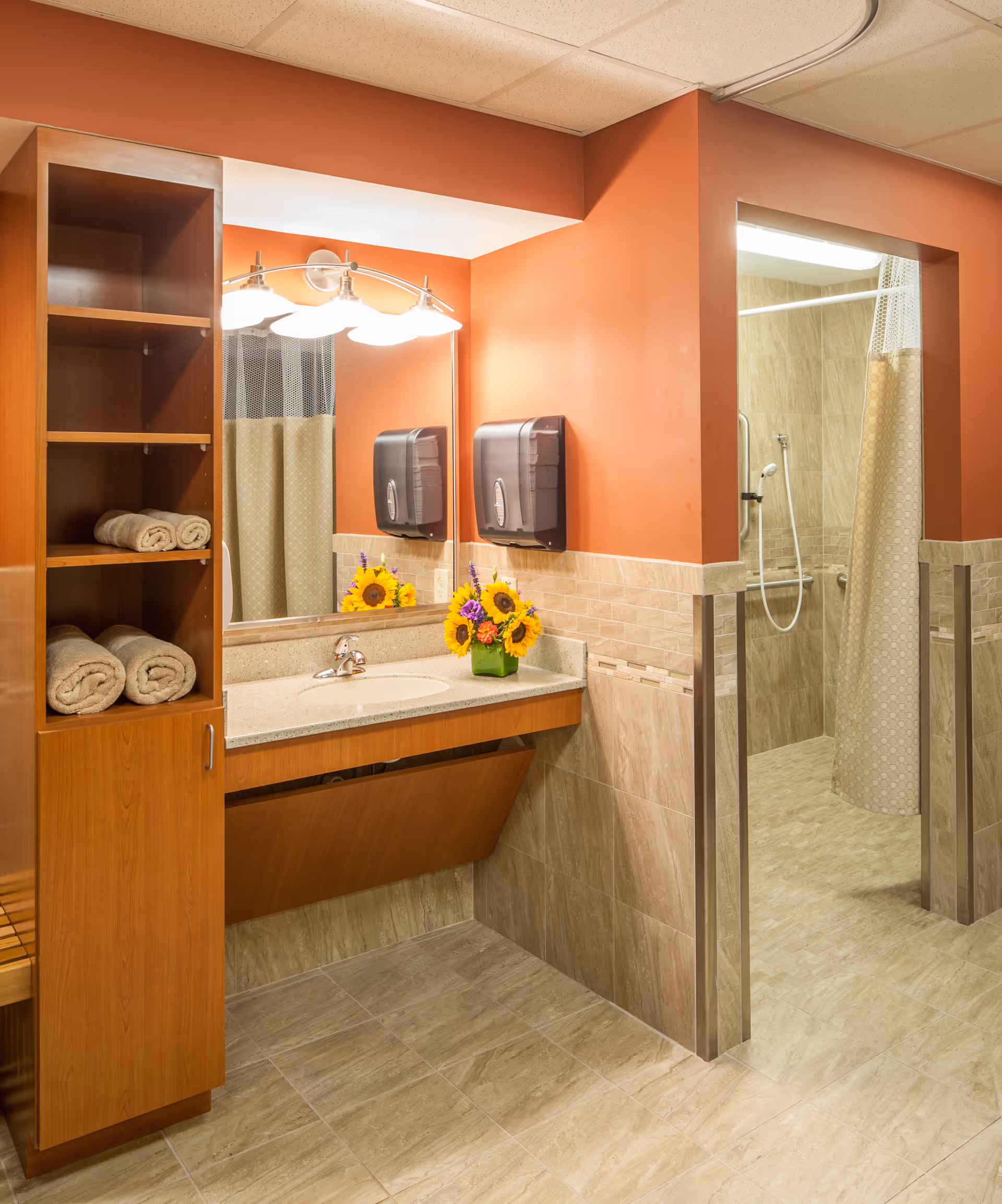 Accessible restroom with a sink and mirror, shelves with rolled towels, and an open tiled shower stall.