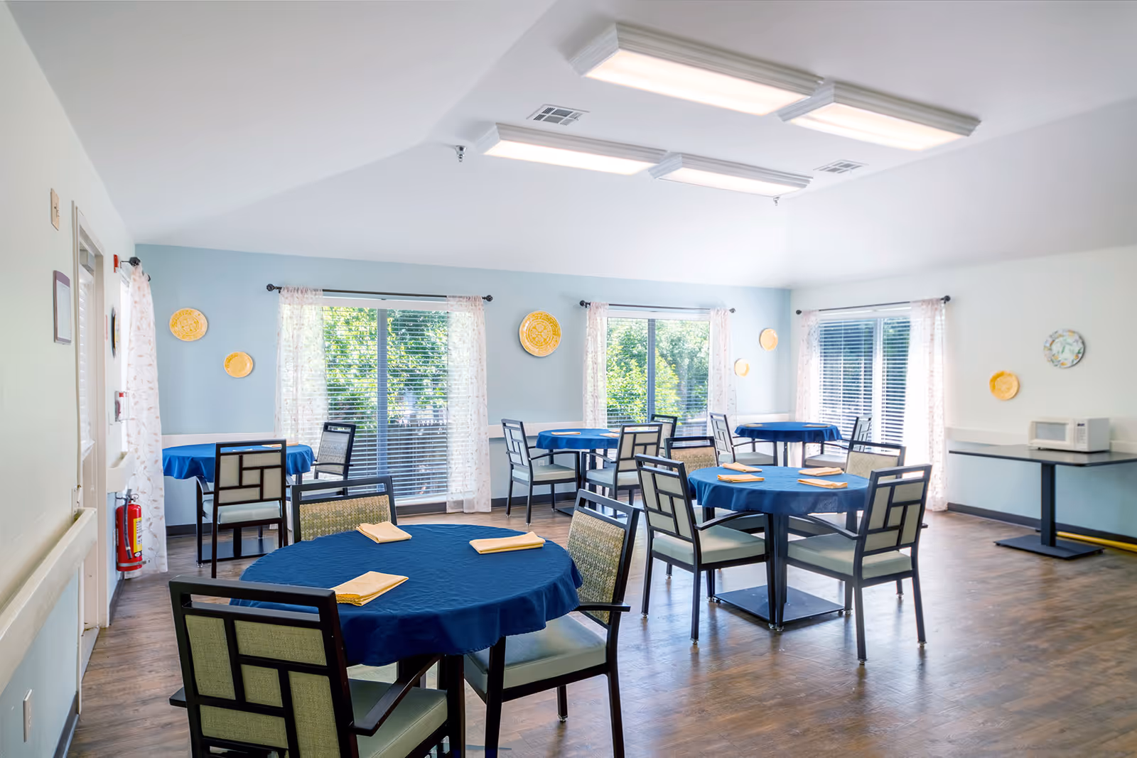 A bright dining room with several round tables covered with blue tablecloths and set with folded yellow napkins. Each table is surrounded by chairs with patterned backs. Large windows with sheer curtains allow natural light to fill the room, and decorative plates hang on the light blue walls. A microwave sits on a table in the corner.