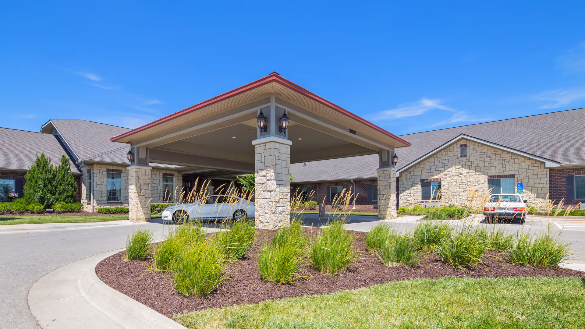Covered entrance porte-cochere and landscaped driveway of a single-story senior living facility under a clear blue sky.