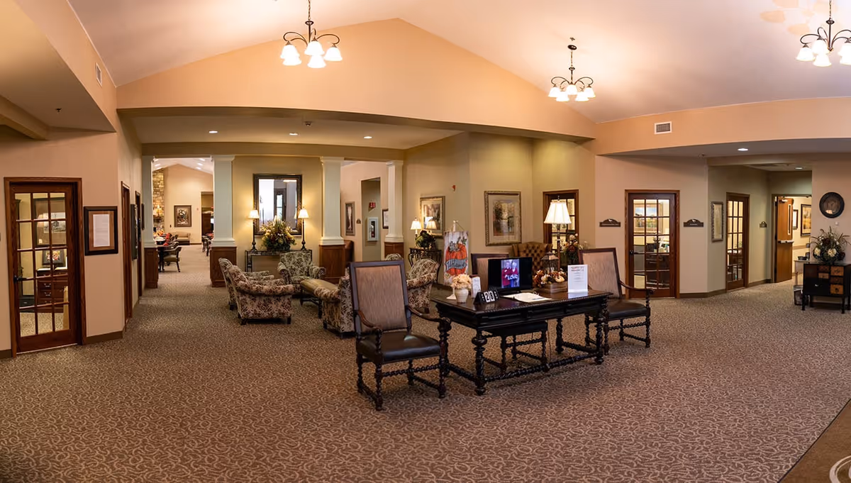 A spacious and warmly lit senior living facility common area with patterned carpet flooring, beige walls, and multiple seating arrangements including upholstered chairs and a wooden table with chairs. The area features decorative lamps, framed artwork on the walls, and overhead light fixtures. Several doorways lead to other rooms, and there is a small TV on the table displaying a program.