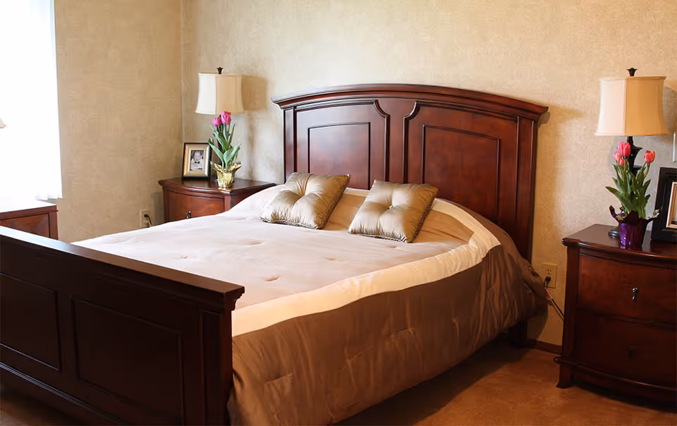 Neatly made bed with a dark wooden headboard flanked by matching nightstands, lamps, and vases of flowers in a warmly decorated bedroom.