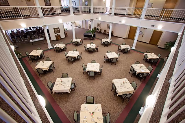 A spacious dining room with multiple square tables covered with beige tablecloths, each set with plates, glasses, and silverware. The room has a high ceiling with a second-floor balcony overlooking the area. There is a black grand piano against the far wall and several chairs arranged in rows in a section to the left. The walls are decorated with framed pictures and the room is well-lit with natural light from windows and ceiling lights.