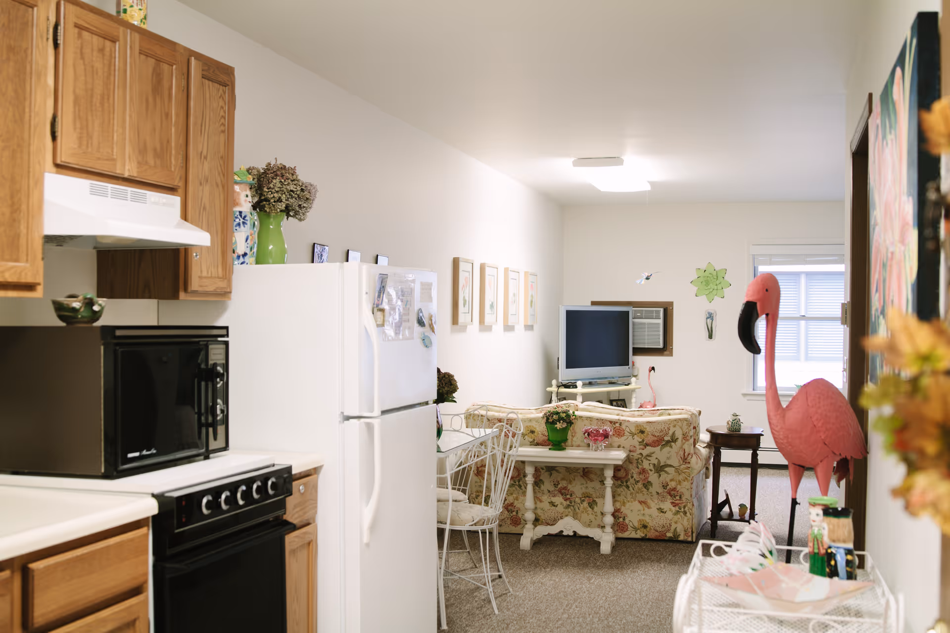 View of a small senior living apartment showing a kitchen area with wooden cabinets, a white refrigerator, a microwave, and a stove on the left. Beyond the kitchen is a living room with a floral-patterned sofa, a small white table with a green plant, a TV on a stand, and a large pink flamingo decoration near the window.