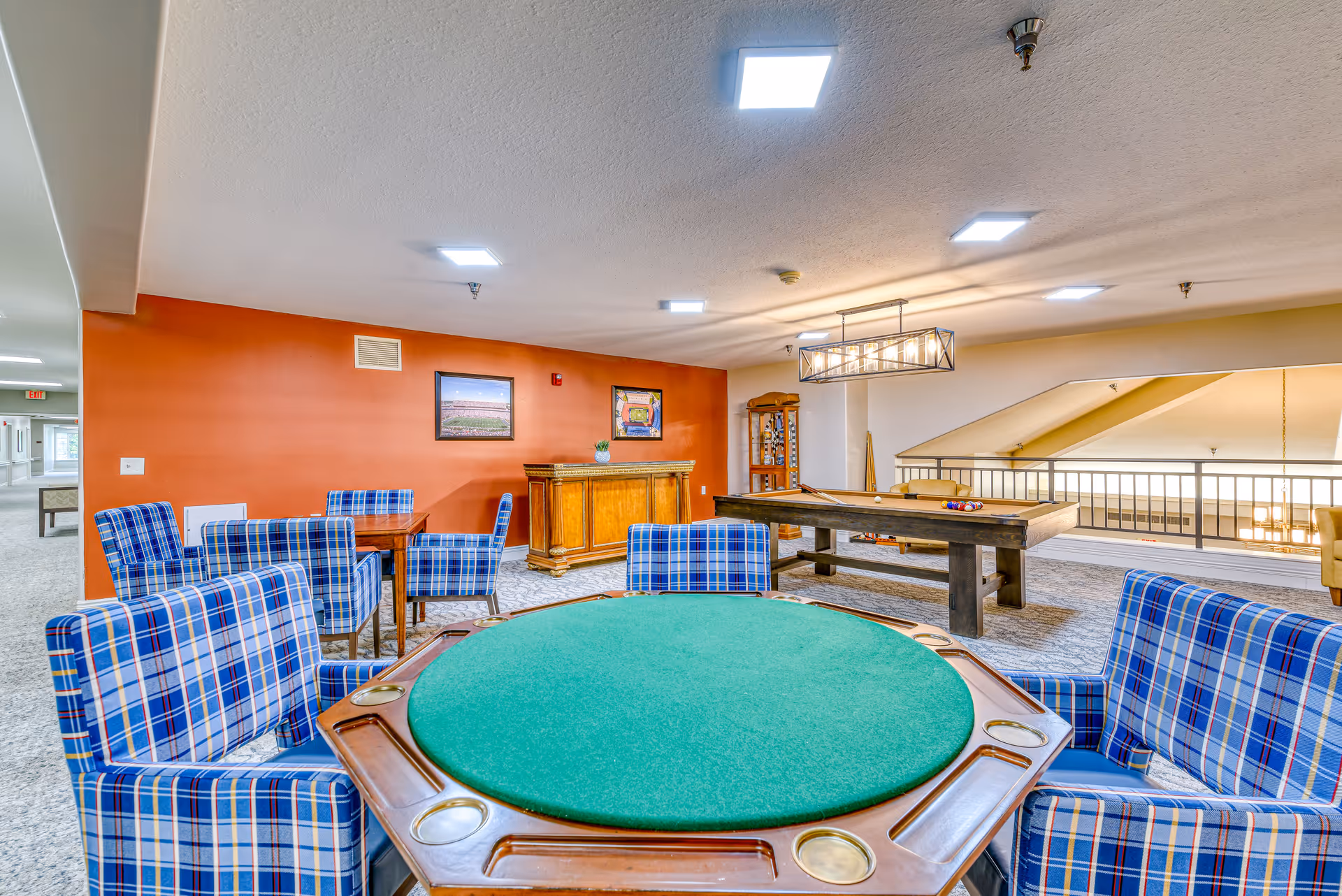 A recreational room in a senior living facility featuring a green felt poker table surrounded by blue plaid upholstered chairs. In the background, there is a pool table with billiard balls arranged, a wooden bar cabinet, and framed pictures on an orange accent wall. The room has bright overhead lighting and a railing overlooking a lower level.