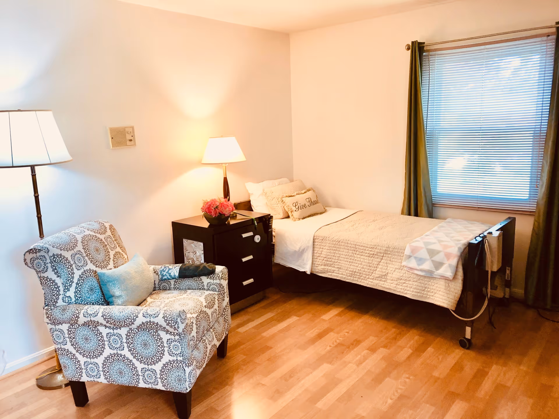 Furnished bedroom with a single bed, patterned armchair, nightstand with a lamp, and a window with blinds.