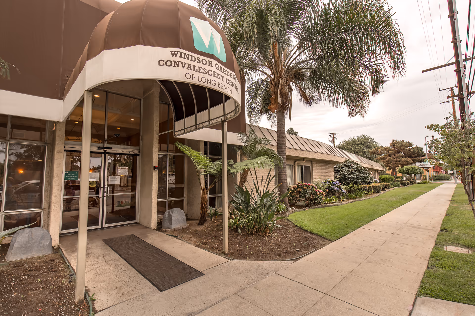 Entrance of Windsor Gardens Convalescent Center of Long Beach with a brown and white canopy above the door, surrounded by palm trees and landscaped greenery along a sidewalk.