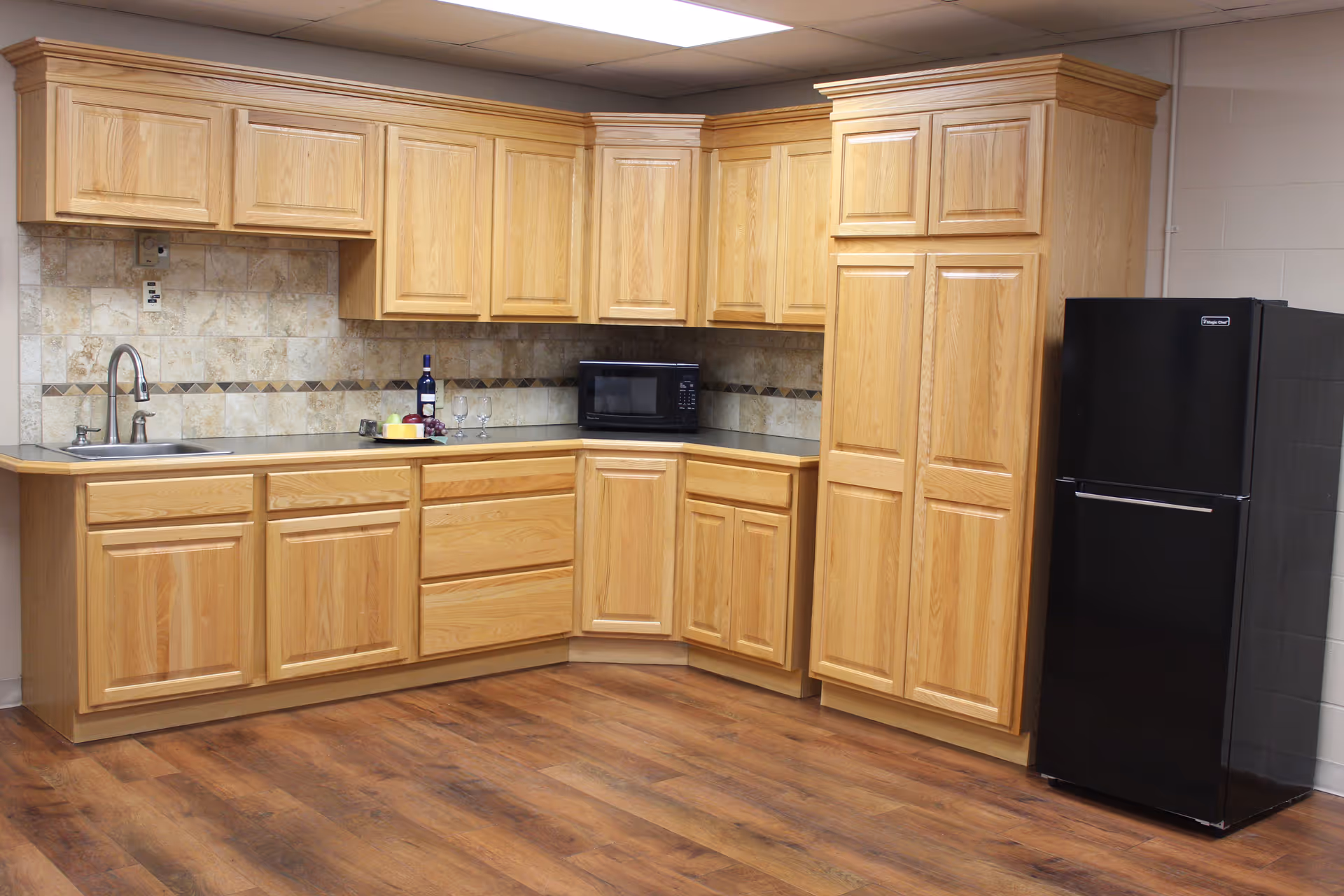 A kitchen area with light wood cabinets, a black refrigerator, a microwave, a sink with a faucet, and a countertop with a bottle of wine, two wine glasses, and a small plate with cheese and grapes. The floor is wooden and the backsplash has a beige tile design.