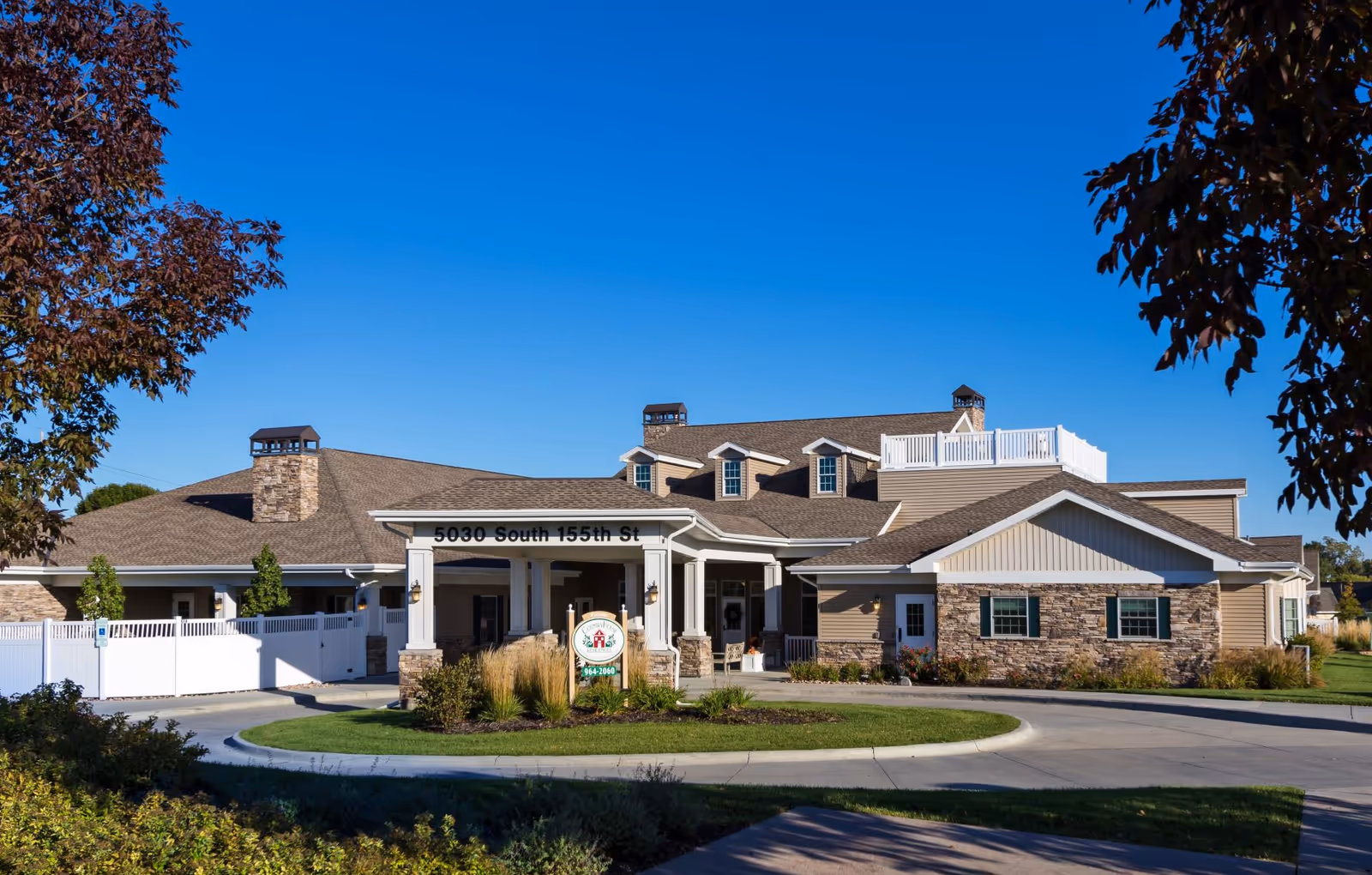 Exterior view of CountryHouse Residence, a single-story building with beige siding and stone accents, a covered entrance with white columns, and a sign displaying the facility's name and phone number. The building is surrounded by landscaped greenery and trees under a clear blue sky.
